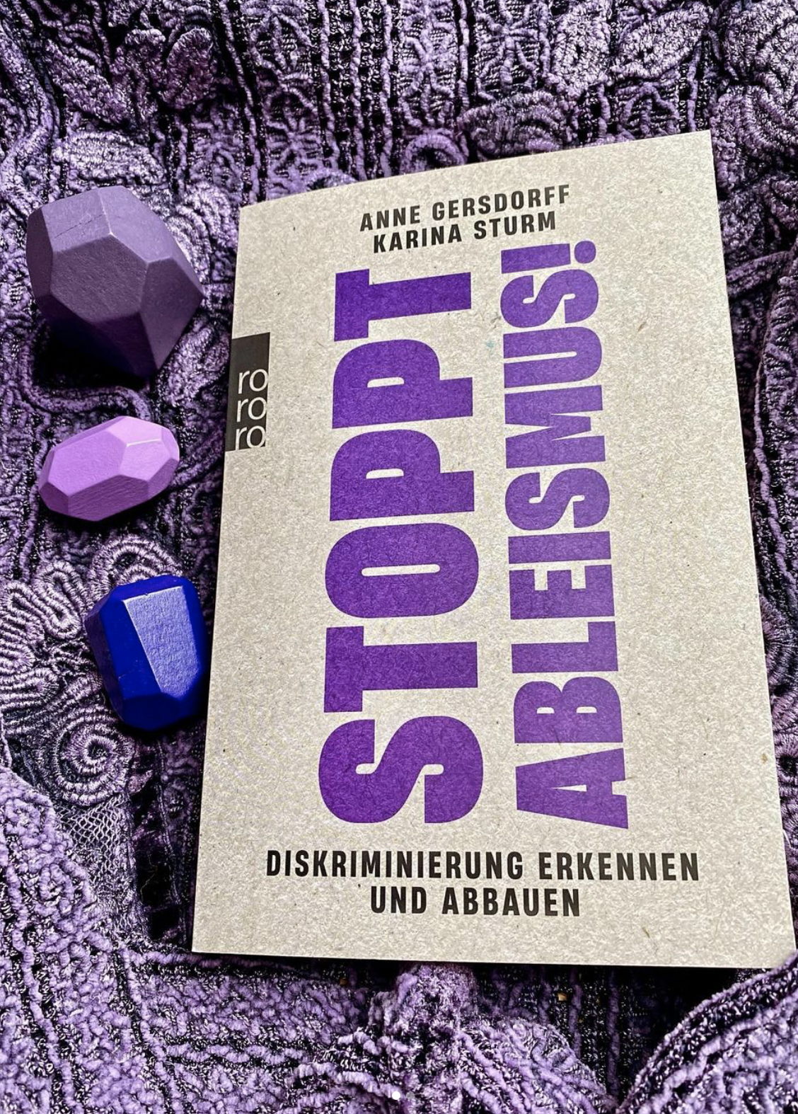 Das Buch Stoppt Ableismus auf einem lilafarbenen Schal mit kleinen lila Holzformen.